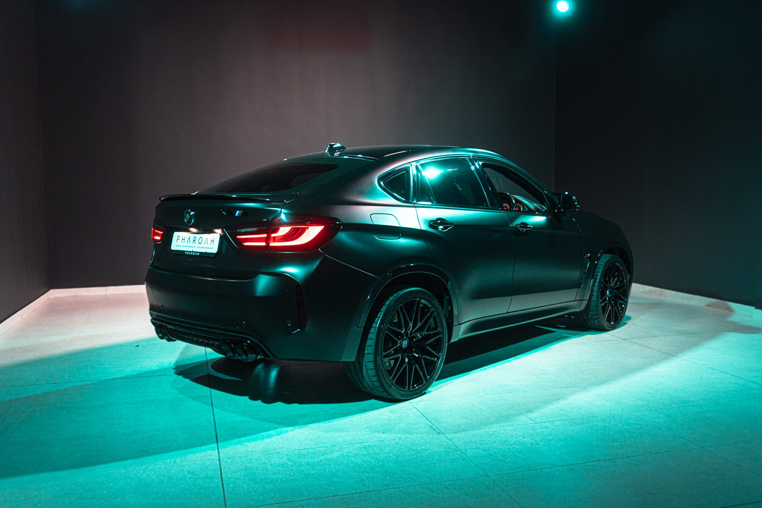 Rear three-quarter view of a dark teal sports sedan illuminated by turquoise showroom lighting in a dim space.