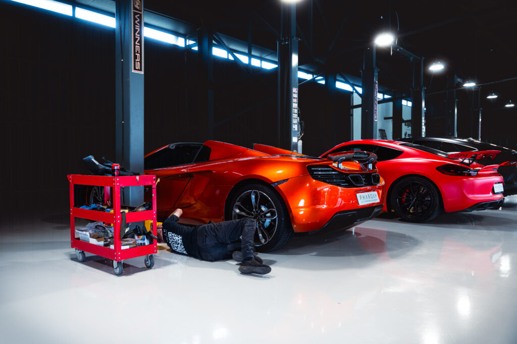 Mechanic lying under a bright red sports car in a modern, blue-lit garage with a red tool cart nearby.