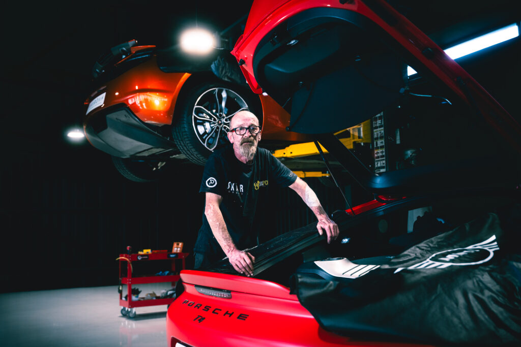 Man with glasses and beard working beneath a red Porsche that’s raised on a hydraulic lift in a dim garage.