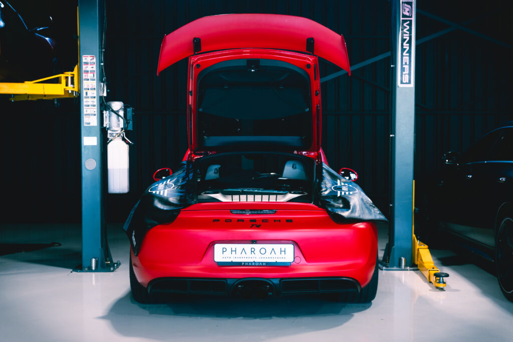 Red Porsche with hood open, positioned on a car lift in a dim workshop, seen from the rear with a visible PHAROH license plate arcada.