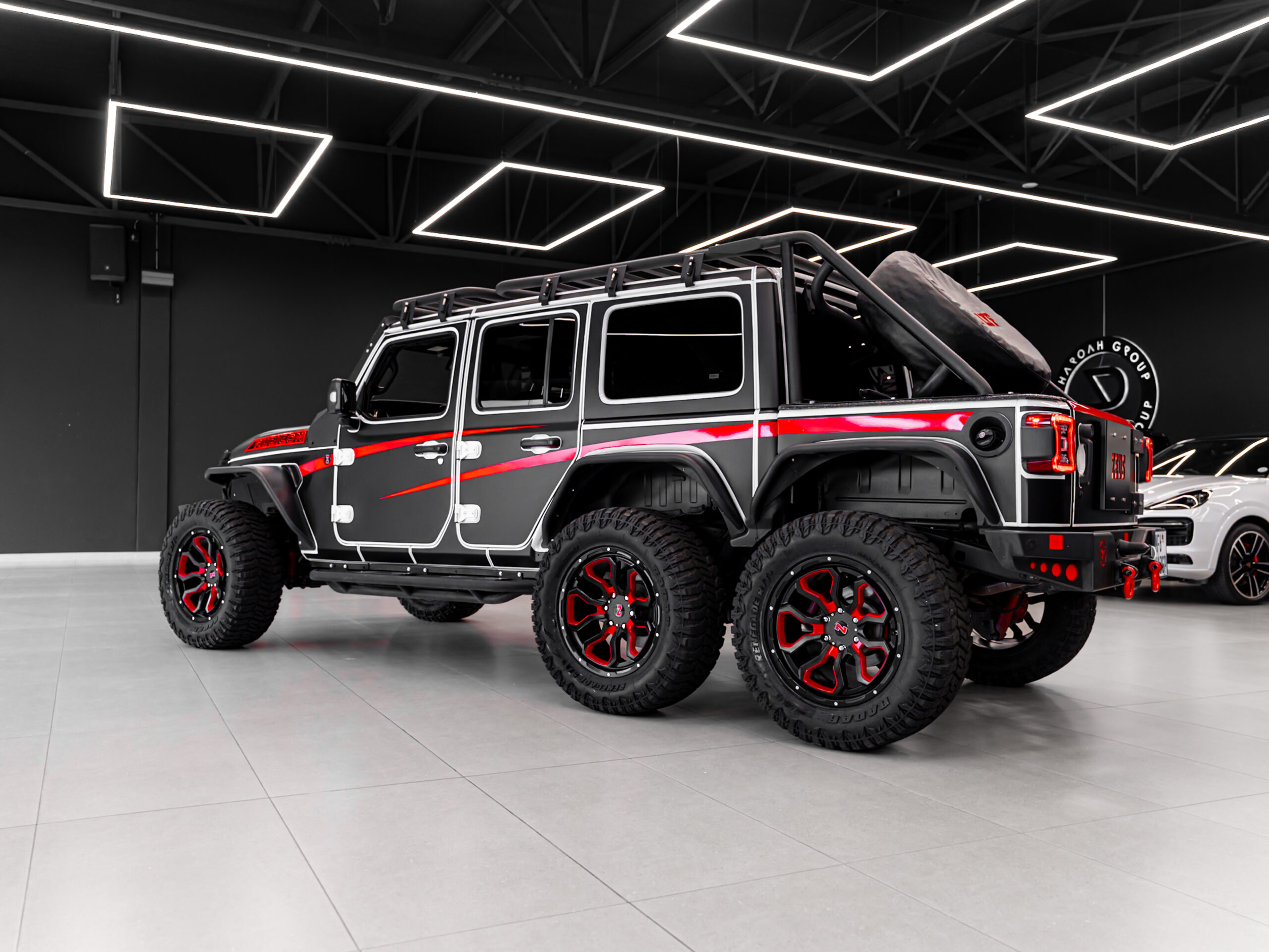 Black and red off-road SUV with large mud-terrain tires in a showroom, featuring a roof rack and spare tire at the rear under bright geometric ceiling lights.