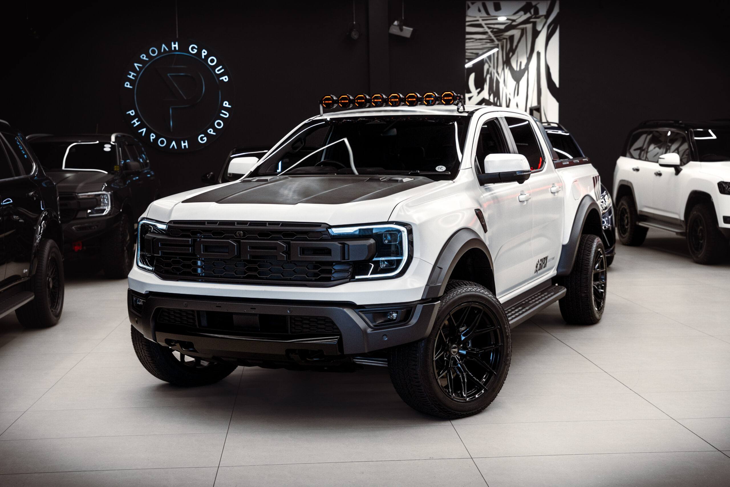 White Ford pickup with an orange roof-mounted light bar in a showroom, with other trucks in the background.