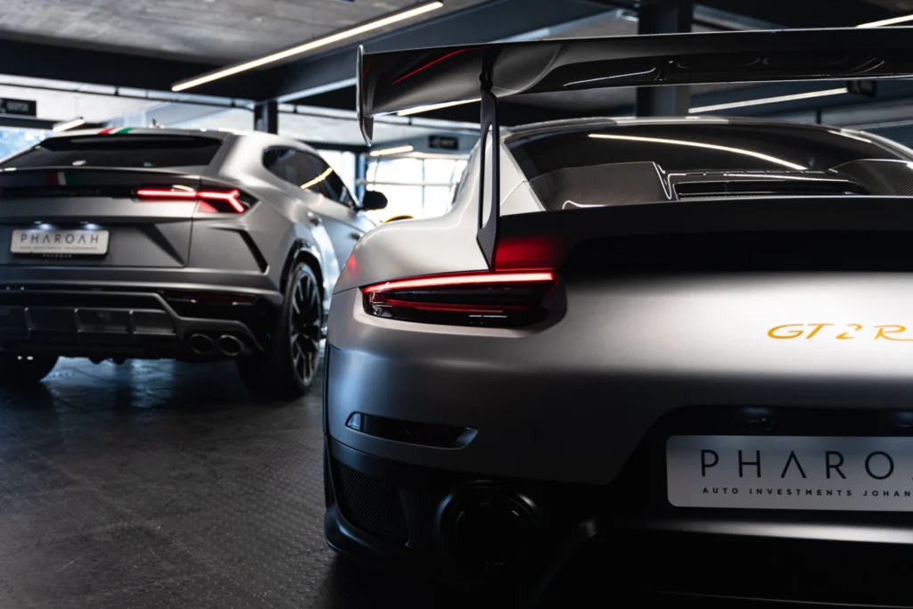 Close-up of a silver Porsche GT2 RS rear with a large carbon-fiber wing and red taillight in a showroom, another car in the background, dim lighting.