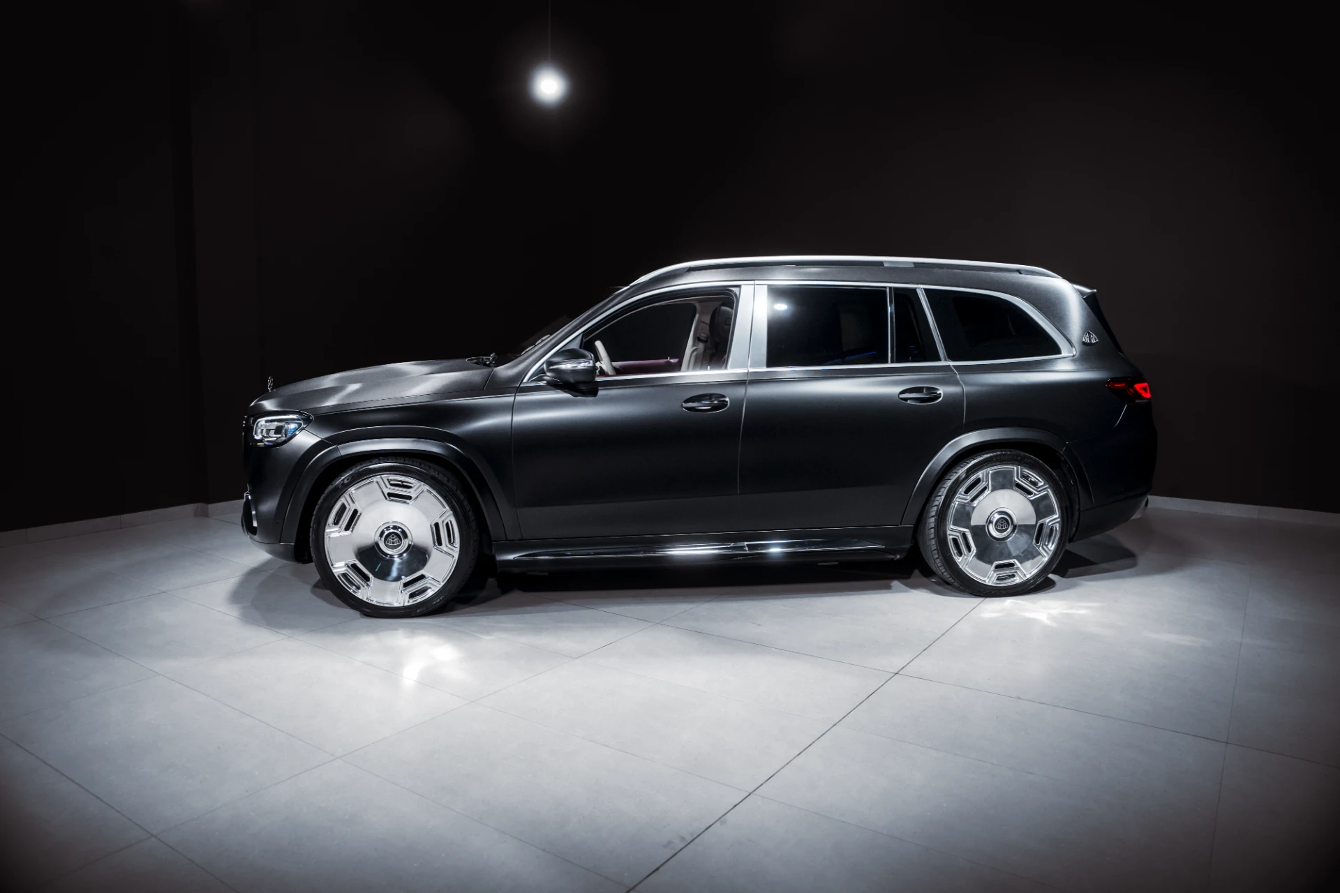Side view of a dark luxury SUV on a showroom floor with bright chrome wheels and a black background.