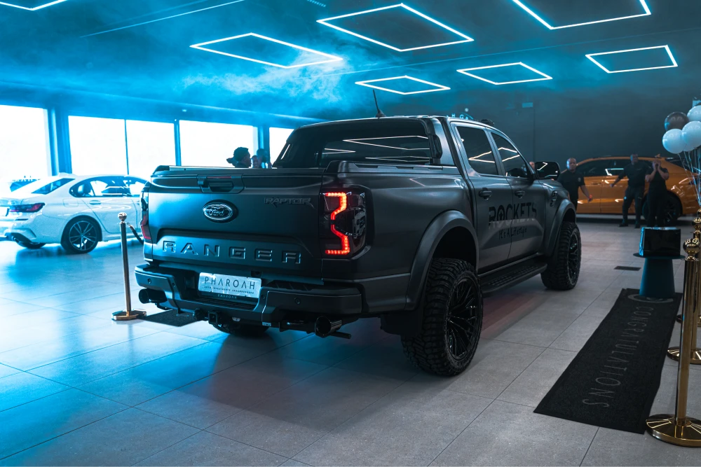 Black Ford Ranger pickup on a showroom floor under blue neon lights, with visitors and other cars in the background.