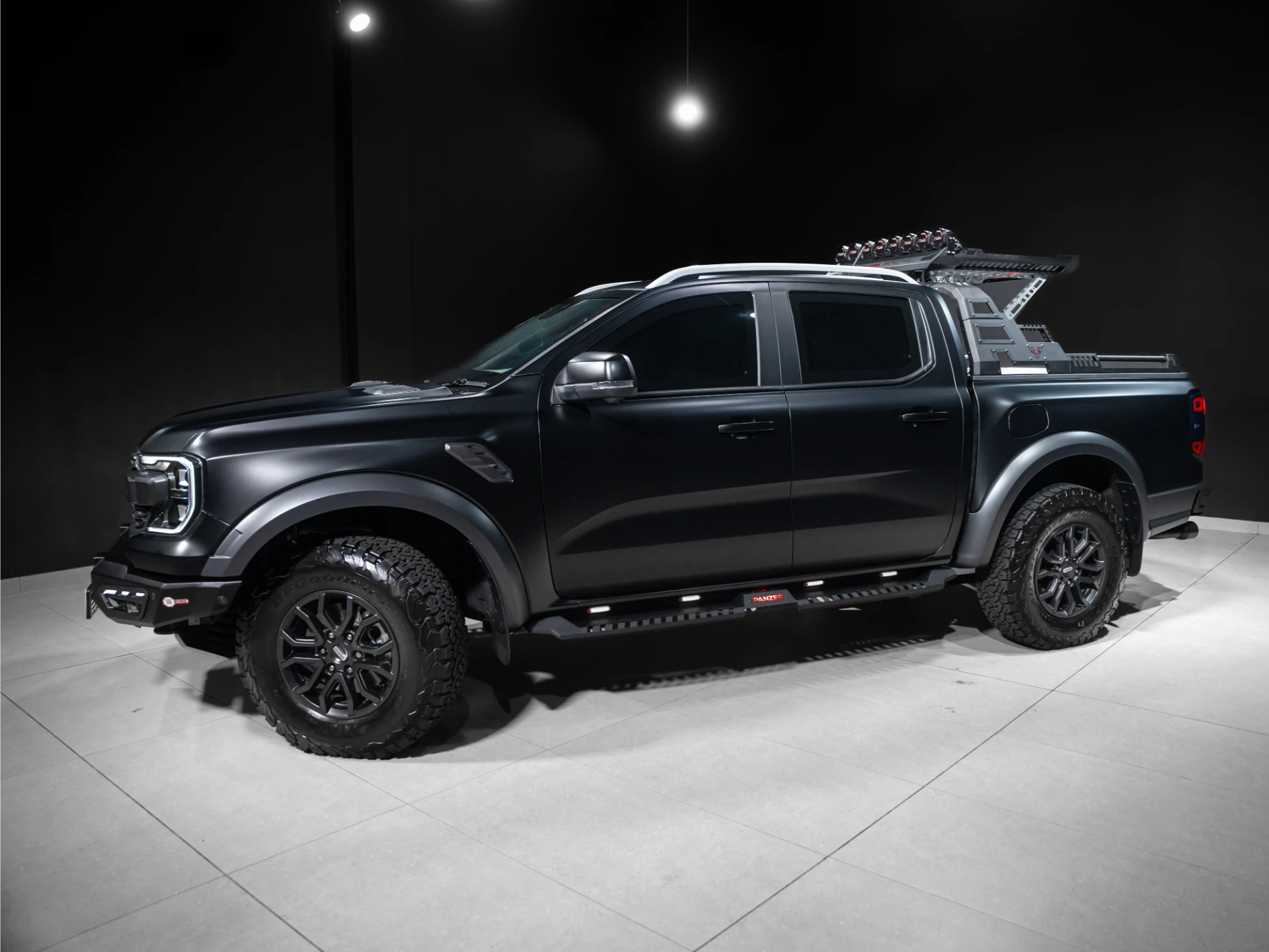 Black pickup truck with large off-road tires and roof rack/ladder system displayed in a dim showroom area.