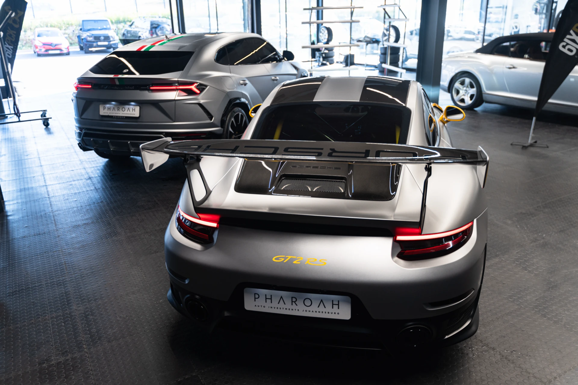 Gray Porsche GT2 RS rear view with large carbon spoiler in a showroom, other sporty cars visible in background with showroom lighting and windows behind.