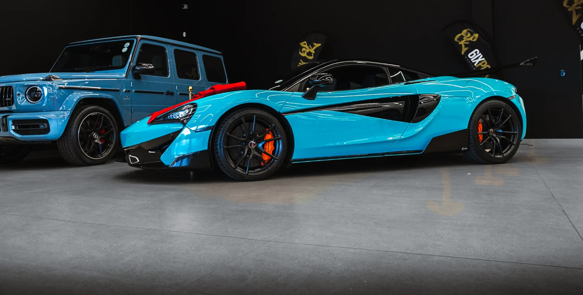 Blue McLaren sports car with black accents parked in a showroom beside a blue Mercedes G-Class.