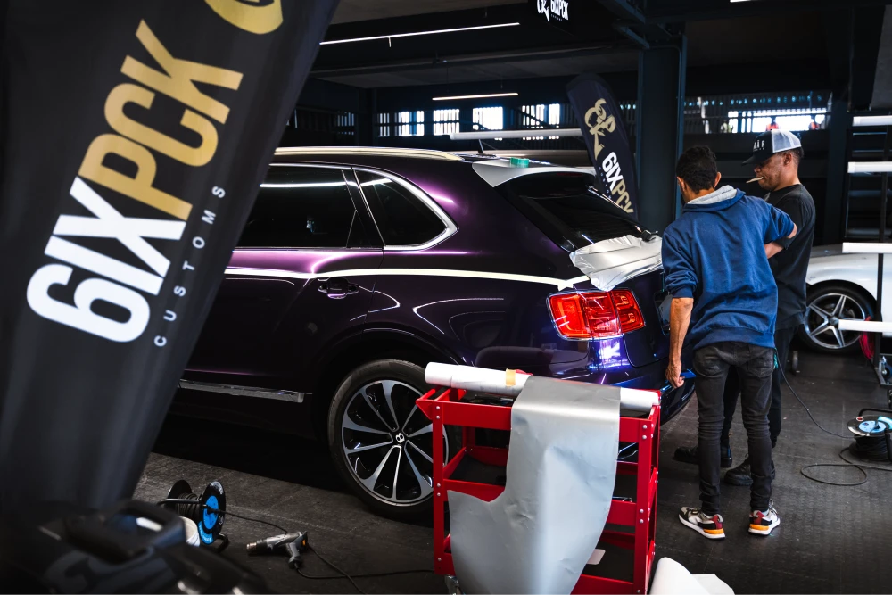 Two technicians stand by the rear of a glossy dark purple SUV in a car custom shop, with a red tool cart and banners in the background.