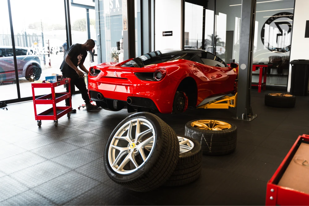 Red sports car on a lift in a car workshop, a mechanic at work nearby with a red tool cart and several tires on the floor in front of it.