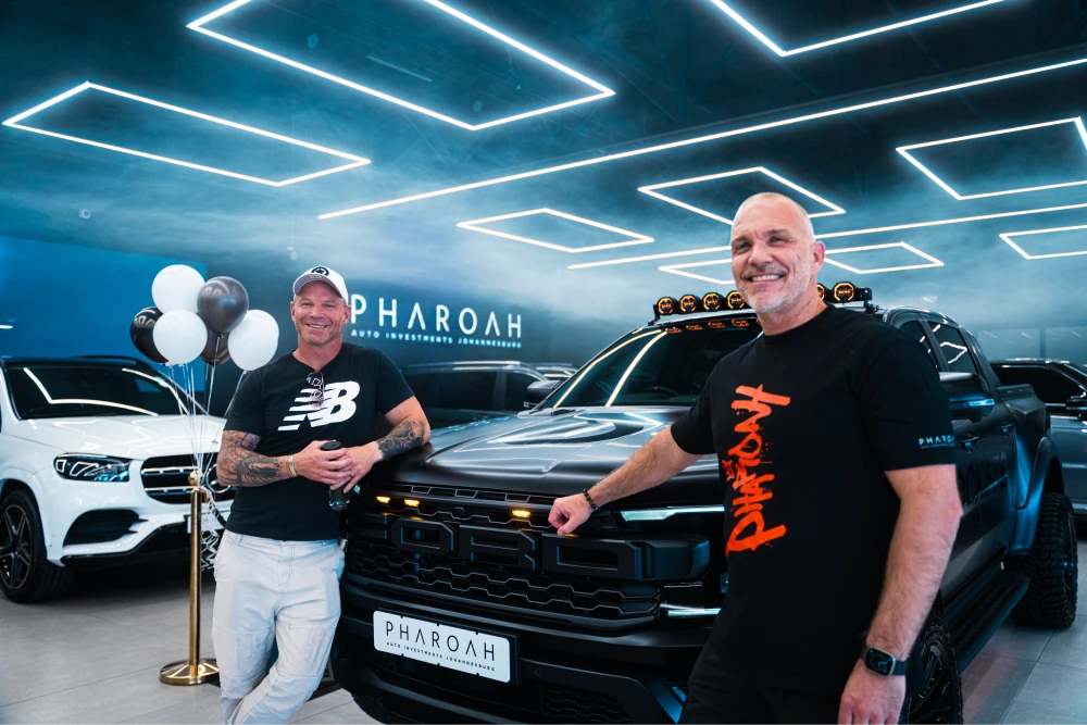 Two men stand beside a black pickup truck in a bright showroom with neon ceiling lights and balloons in the background, smiling at the camera.