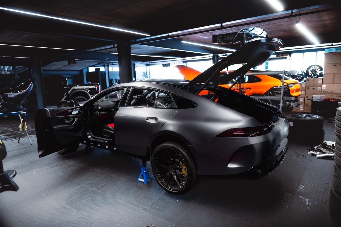Matte gray sports coupe on a blue jack stand in a dim garage, doors and rear hatch open, with tools and tires around.