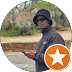 Person wearing a hat, sunglasses, and dark jacket on a tree-lined path outdoors, with an orange star badge in the lower-right corner.